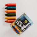 A set of six natural beeswax bath crayons in assorted colors, displayed next to their packaging which is labeled 'Bath Time Crayons'. The packaging is recyclable kraft material with a blue accent and the crayons are in a line on a white background.